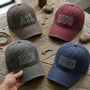 May include: Four baseball caps in various colors: brown, burgundy, gray, and navy blue. Each cap has a different three-letter city abbreviation embroidered in white block letters: ATL, HUS, DEN, and ORD. The caps are displayed on a wooden surface.
