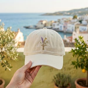 May include: A beige baseball cap with a curved brim, featuring an embroidered floral design of lavender and yellow flowers. The cap is held up against a scenic background of a coastal town and the sea.