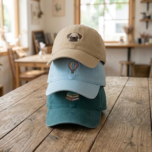 May include: A stack of three baseball caps in tan, light blue, and teal. The tan cap features a pug face, the light blue cap has a hot air balloon, and the teal cap has a stack of books embroidered on the front. The caps are on a wooden surface.