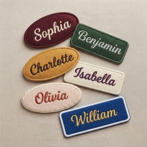 May include: A collection of six felt name patches in various shapes and colors. The patches feature embroidered names including Sophia, Benjamin, Charlotte, Isabella, Olivia, and William in different thread colors and script or print fonts.