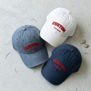 May include: Three baseball caps in white, grey, and navy blue. Each cap features the word CUSTOM embroidered in red capital letters, with the words YOUR DESIGN embroidered in smaller red letters directly underneath the main text on the front.