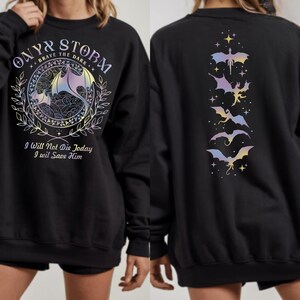 The image is a collage of two photos of a black sweatshirt with a dragon design and text. The back of the sweatshirt has a design of bats and stars, and the front has a design of a dragon and text.