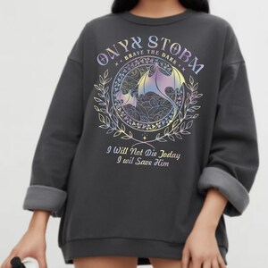 a person wearing a grey sweatshirt with a graphic design on it. The design includes a dragon and a floral pattern, along with the text "ONYX STORM" and "I WILL NOT Die Today I WILL SAVE HIM".