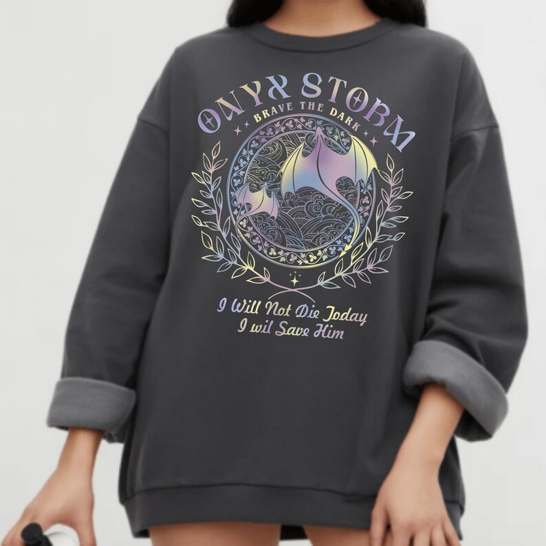 a person wearing a grey sweatshirt with a graphic design on it. The design includes a dragon and a floral pattern, along with the text "ONYX STORM" and "I WILL NOT Die Today I WILL SAVE HIM".