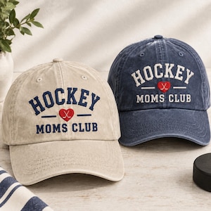 May include: Two baseball caps, one beige and one navy blue, both featuring embroidered text that reads HOCKEY MOMS CLUB with a red heart containing two crossed hockey sticks in the center. The caps have a distressed, vintage-style fabric finish.