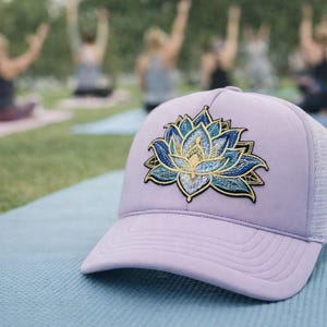 Puede incluir: Gorra de béisbol lavanda con la parte trasera de malla blanca. La gorra presenta un diseño de flor de loto bordada en tonos azules y dorados. El sombrero está sobre una esterilla de yoga azul, con gente practicando yoga al fondo.