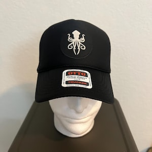 Könnte beinhalten: Schwarze Trucker-Mütze mit einem weißen Tintenfisch-Emblem auf der Vorderseite. Die Mütze hat einen gebogenen Schirm und eine Mesh-Rückseite. Das Etikett lautet "OTTO" und "Trucker".