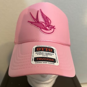Puede incluir: Gorra rosa tipo trucker con un diseño de pájaro bordado en rosa. La gorra presenta una parte delantera de espuma y una banda para el sudor de rizo. Cierre a presión duradero y texto "OTTO WORLD FAMOUS FOAM FRONT TRUCKER OSFM".