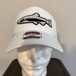 May include: A white trucker hat featuring a black and white embroidered fish patch on the front. The hat includes a sticker on the brim with the text OTTO WORLD FAMOUS FOAM FRONT OSFM TRUCKER. The cap has a structured foam front and a curved brim.