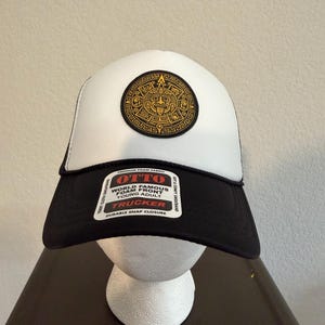 May include: A white and black trucker hat featuring a circular gold and black Aztec calendar patch on the front. A rectangular sticker on the black brim displays the text OTTO WORLD FAMOUS FOAM FRONT YOUNG ADULT TRUCKER DURABLE SNAP CLOSURE.