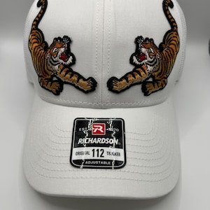 May include: White Richardson 112 Trucker hat with two embroidered tigers in orange, black, and tan. The tigers are positioned on the front panels. The hat has a curved brim and an adjustable strap.