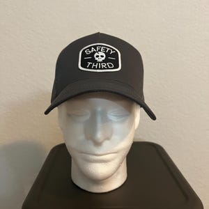 Może przedstawiać: Ciemnoszara czapka typu trucker z zakrzywionym daszkiem. Czapka posiada białą naszywkę z napisem "SAFETY THIRD" i grafiką czaszki. Czapka jest prezentowana na białej głowie manekina.