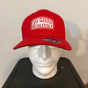 Op de afbeelding: Een rode baseballcap met een witte mesh achterkant. De voorkant van de cap heeft de tekst "DO GOOD RECKLESSLY" in een witte rechthoekige patch. De cap heeft een gebogen klep en een zwarte onderkant.