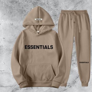 Peut inclure: Un ensemble de sweat à capuche et de pantalon de survêtement beiges. Le sweat à capuche porte le mot "ESSENTIALS" en grandes lettres noires sur le devant et une petite étiquette sur la capuche. Le pantalon de survêtement a une taille à cordon et le mot "ESSENTIALS" sur la jambe.