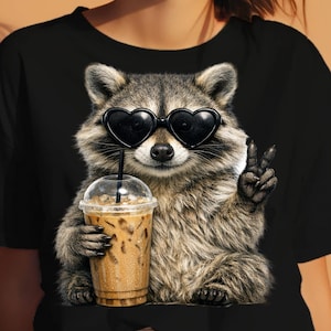 Puede incluir: Camiseta negra con un mapache con gafas de sol en forma de corazón y un café helado. El mapache hace el signo de la paz. Un diseño divertido y caprichoso, ideal para el uso diario.