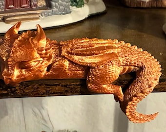 Puede incluir: Figura de dragón naranja con escamas, cuernos y alas detalladas, tumbada sobre una superficie de madera. La figura es un objeto decorativo de temática fantástica.