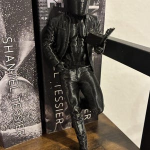Könnte beinhalten: Eine schwarze Figur einer Person mit Helm und Jacke, die sich an zwei Bücher lehnt. Die Figur hält ein Buch und trägt eine Jacke, Hose und Stiefel. Die Bücher haben weißen Text auf schwarzem Hintergrund.
