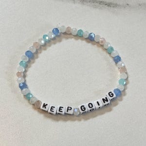 May include: A beaded bracelet featuring small, faceted, translucent beads in shades of light blue, white, and pale pink. The bracelet includes white square letter beads that spell out the phrase KEEP GOING in black capital letters.