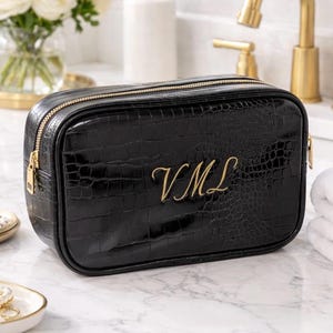 Könnte beinhalten: Schwarze rechteckige Kosmetiktasche mit Krokodiltextur und goldenem Reißverschluss. Die Tasche hat goldene Initialen "VML" auf der Vorderseite. Die Tasche steht auf einer Marmoroberfläche.