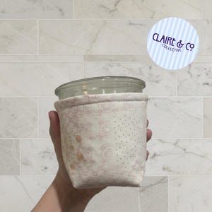 May include: A fabric jar cosy with a floral pattern in shades of pink and cream. The cosy is designed to fit around a glass jar. The image also includes a round logo with the text "Claire & Co Collective".