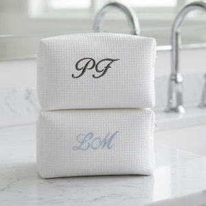 May include: Two white, textured cosmetic bags stacked on a marble surface. The top bag has the embroidered initials "PF" in black, while the bottom bag features "LM" in light blue. The bags are in a bathroom setting.