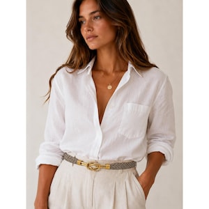 May include: A white linen shirt with a chest pocket and long sleeves. The shirt is paired with beige pants and a gray and gold belt. A gold necklace with a round pendant is also visible. The outfit is simple and elegant.