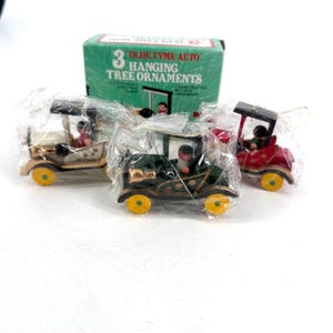 Peut inclure: Trois décorations de sapin de Noël Olde Tyme Auto dans une boîte. Les ornements sont des voitures miniatures en bois dorées, vertes et rouges, chacune avec une petite figurine à l'intérieur. Les voitures ont des roues jaunes et sont emballées dans du plastique transparent.