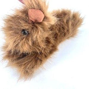 May include: A brown plush hand puppet featuring long, shaggy synthetic fur, a single visible black plastic eye, and a small, rounded tan fabric ear. The puppet has an open mouth design, allowing for manual manipulation of the jaw area.