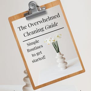 May include: A wooden clipboard with a silver clip holds a white paper with the title "The Overwhelmed Cleaning Guide". The paper also includes the text "Simple Routines to get started!". A white vase with white flowers sits on a white surface. The text "THIS IS A DIGITAL DOWNLOAD" is at the bottom.