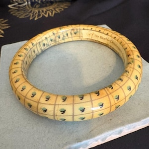 May include: A cream-colored bangle bracelet with a grid pattern and small green accents. The bracelet has a smooth, glossy finish and the text "for Buchi-Dechmann Co." is visible. The bracelet is resting on a light blue surface.