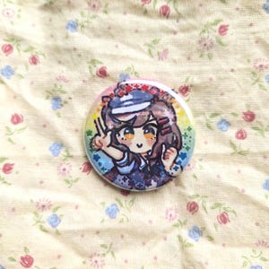 K&ouml;nnte beinhalten: Ein runder Ansteckbutton mit einer Anime-Illustration eines M&auml;dchens in Matrosenkleidung. Der Button hat einen Regenbogenrand und liegt auf einem Stoff mit Blumenmuster.