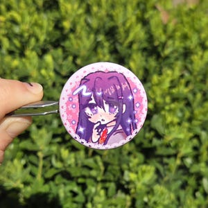 May include: A round button pin featuring an anime-style illustration of a girl with purple hair and eyes. The pin has a pink border with hearts and sparkles. The background is a blurred green.