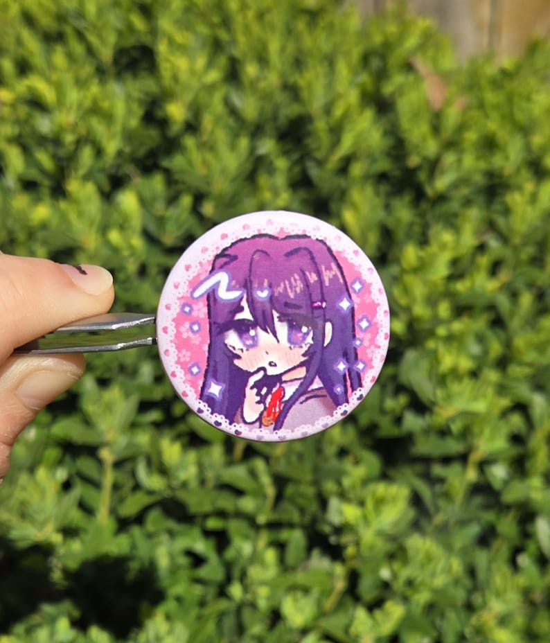 May include: A round button pin featuring an anime-style illustration of a girl with purple hair and eyes. The pin has a pink border with hearts and sparkles. The background is a blurred green.