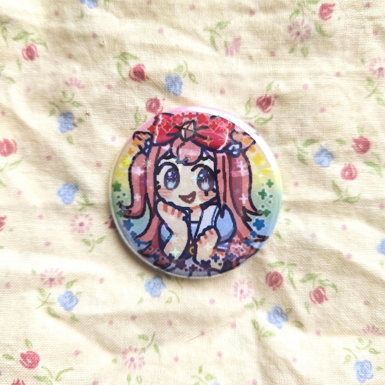 K&ouml;nnte beinhalten: Ein runder Button mit einer farbenfrohen Anime-Illustration eines M&auml;dchens mit rosa Haaren, einer roten Schleife und einem blauen Kleid. Der Button hat einen Regenbogenrand und ist vor einem floralen Hintergrund platziert.