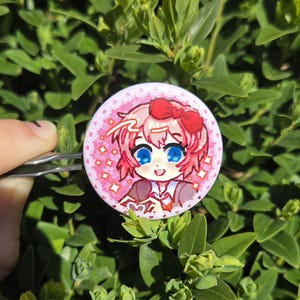 May include: A round, pink button with a cartoon illustration of a character with pink hair, a red bow, and blue eyes. The button has a pink background with white stars and a white border. The button is held by a silver clip against a backdrop of green leaves.