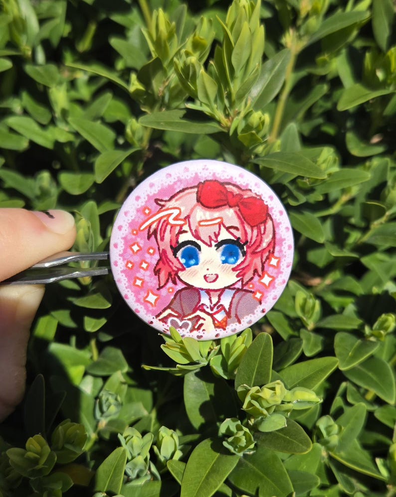 May include: A round, pink button with a cartoon illustration of a character with pink hair, a red bow, and blue eyes. The button has a pink background with white stars and a white border. The button is held by a silver clip against a backdrop of green leaves.