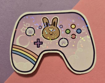 May include: A sticker in the shape of a video game controller. The controller is light purple with a brown bunny face in the center. It has colorful buttons, a rainbow, and paw prints. Glitter accents the design.