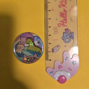 May include: A round button pin with a cartoon illustration of a dinosaur, bunny, and cat. The pin has a holographic effect and is next to a ruler with Hello Kitty branding. The button pin is approximately 1 inch in diameter.