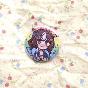 K&ouml;nnte beinhalten: Ein runder Ansteckbutton mit einer farbenfrohen Illustration einer Anime-Figur mit braunem Haar, einem wei&szlig;en Streifen und blauen Akzenten. Die Figur hat einen sch&uuml;chternen Ausdruck und h&auml;lt die H&auml;nde vor einem regenbogenfarbenen Hintergrund zusammen.