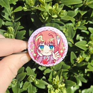 May include: A round, pink button featuring a cartoon character with pink hair, blue eyes, and a red heart. The button has a white lace border and is held against a backdrop of green leaves.