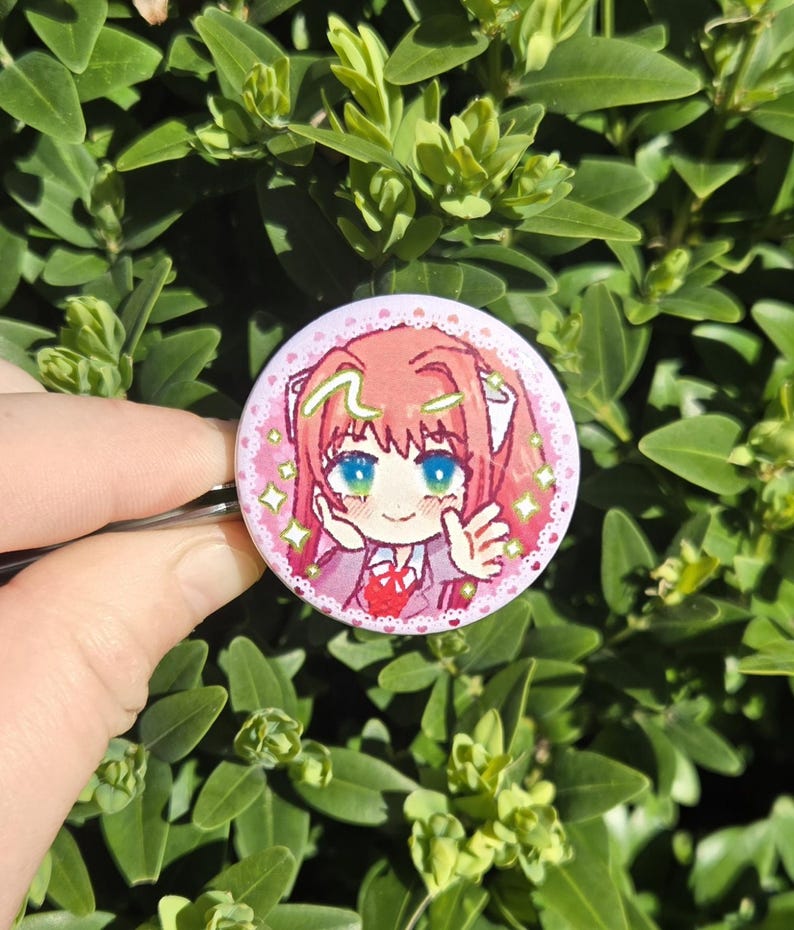 May include: A round, pink button featuring a cartoon character with pink hair, blue eyes, and a red heart. The button has a white lace border and is held against a backdrop of green leaves.