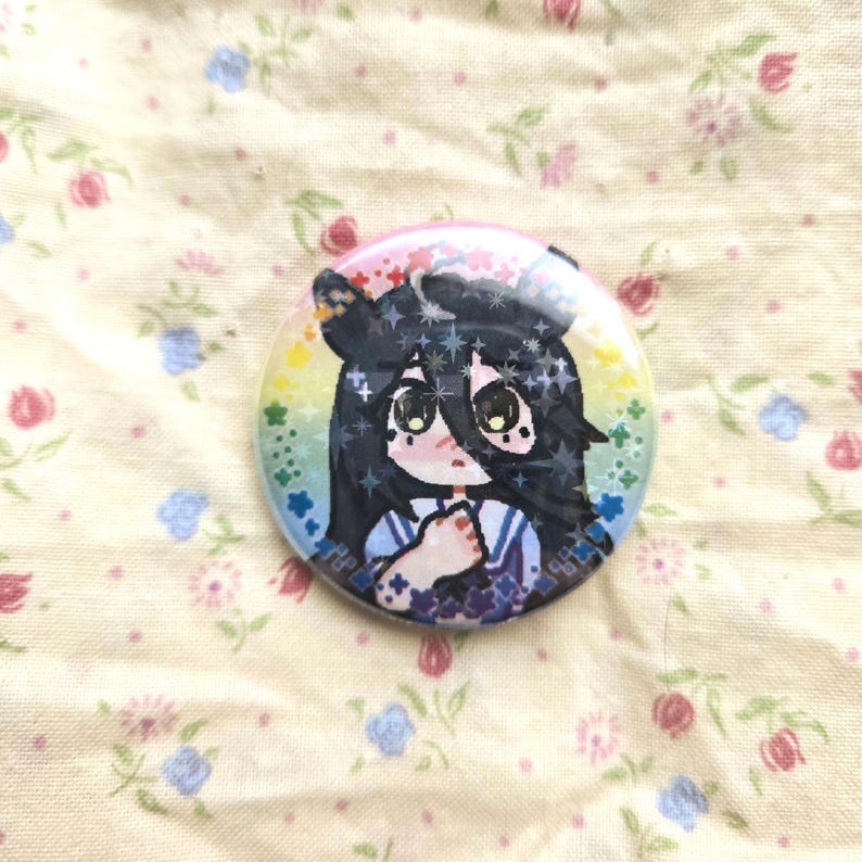 K&ouml;nnte beinhalten: Ein runder Ansteckbutton mit einer holografischen Illustration einer Figur mit langem schwarzem Haar und gro&szlig;en Augen. Die Figur ist vor einem regenbogenfarbenen Hintergrund mit einem sternf&ouml;rmigen Glitzereffekt dargestellt.