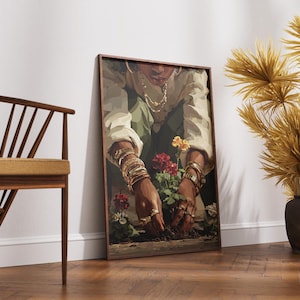 May include: A framed art print depicts a person tending to flowers. The person is wearing gold jewelry and a white shirt. The artwork is leaning against a white wall. A wooden chair and decorative plants are also in the image.