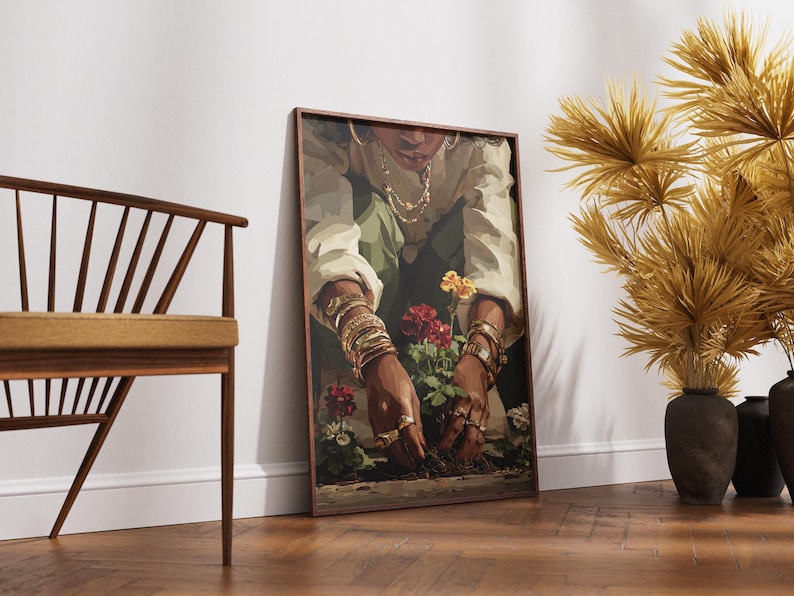 May include: A framed art print depicts a person tending to flowers. The person is wearing gold jewelry and a white shirt. The artwork is leaning against a white wall. A wooden chair and decorative plants are also in the image.