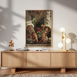 May include: A framed art print depicts a person tending to red flowers. The person is wearing gold jewelry, including bracelets and rings. The artwork hangs above a light wood cabinet with a gold lamp and a wooden chair.