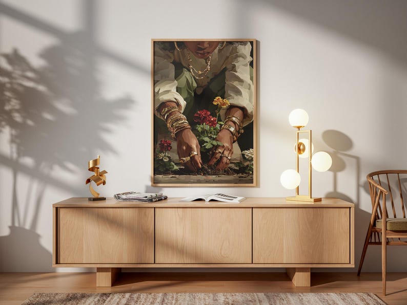 May include: A framed art print depicts a person tending to red flowers. The person is wearing gold jewelry, including bracelets and rings. The artwork hangs above a light wood cabinet with a gold lamp and a wooden chair.