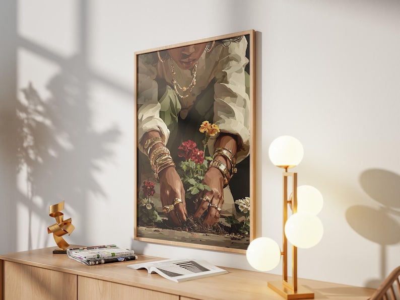 May include: A framed art print depicts a person tending to flowers, adorned with gold jewelry. The artwork is set against a neutral wall, complemented by a modern lamp with spherical lights and a wooden cabinet. A gold sculpture and magazines are also on the cabinet.