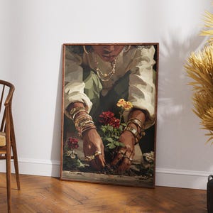 May include: A framed art print depicts a person tending to flowers. The person is wearing gold jewelry and a white shirt. The flowers are red, yellow, and white. The artwork is displayed on a white wall next to a wooden chair and dried plants.