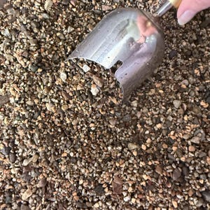 May include: A metal scoop with a light-colored wooden handle is partially submerged in a pile of small, multi-colored gravel. The stones are a mix of tan, brown, gray, and white pebbles of varying shapes and sizes, creating a textured surface.
