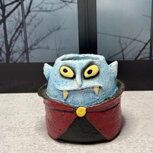 May include: A small, textured planter shaped like a cartoon vampire head with light blue skin, yellow eyes, and two white fangs. The base is designed to look like a red and black cape with a single yellow button at the center of the collar.